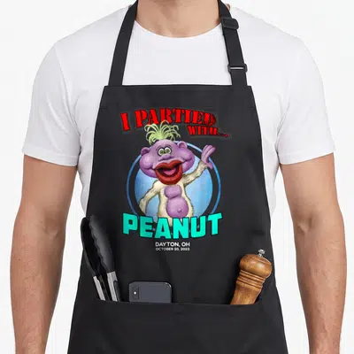 Peanut dayton apron - Purple monster waving inside blue circle with red and cyan text reading I Partied With Peanut — shown on a black cotton canvas apron against a white background