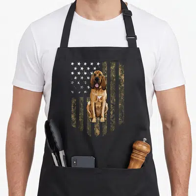 Bloodhound 4th of july apron - Sitting brown bloodhound in front of distressed camo flag with white stars — shown on a black cotton canvas apron against a white background