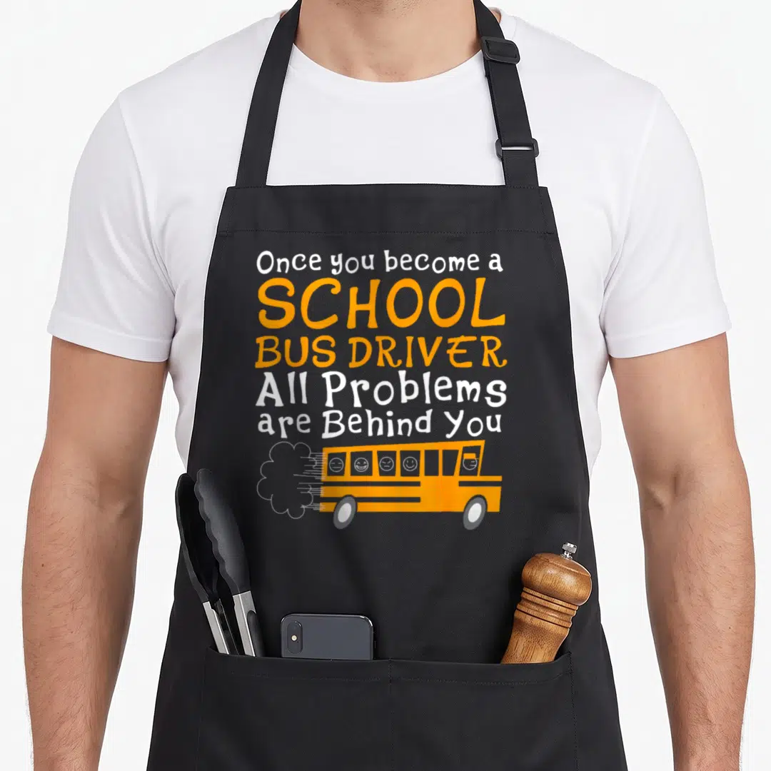 Funny driver school bus apron - Orange cartoon bus facing right with stacked text and four face icons in windows — shown on a black cotton canvas apron against a white background