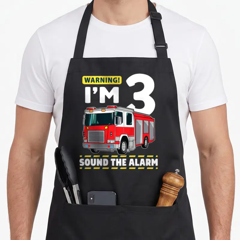 Firefighter 3rd truck apron - Red fire truck with WARNING I'M 3 text and striped borders — shown on a black cotton canvas apron against a white background