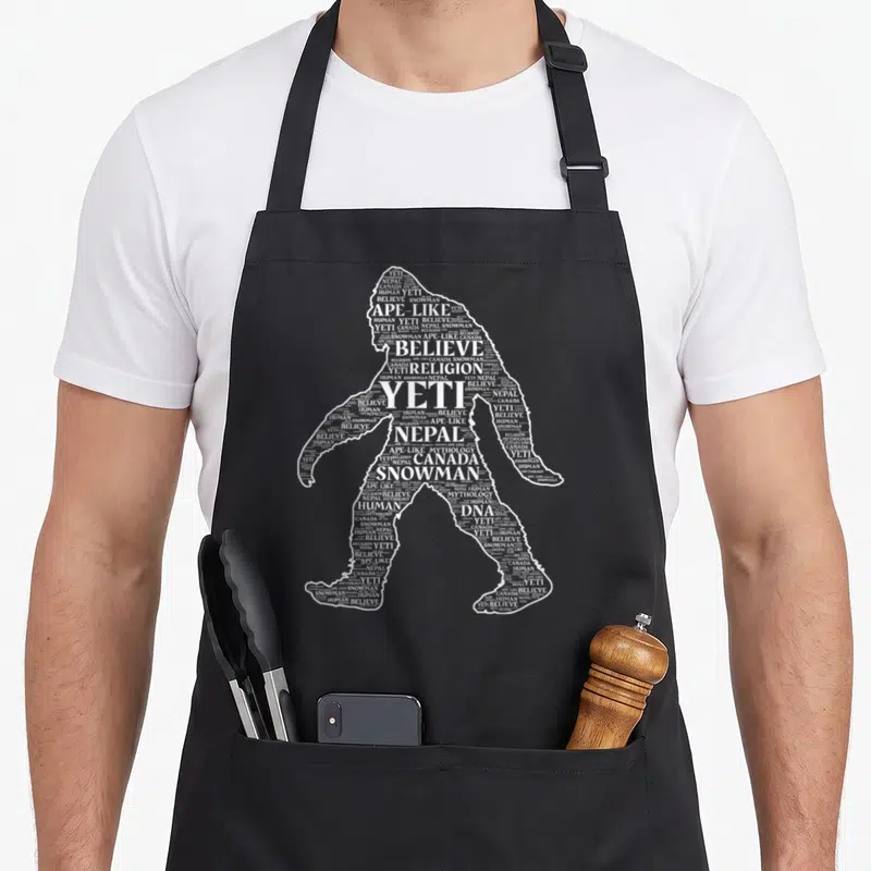 Cloud yeti apron - Walking yeti silhouette formed by white text reading Nepal, Canada, Believe, Ape-Like, Mythology — shown on a black cotton canvas apron against a white background