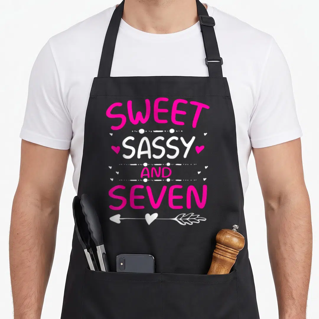 7th sassy apron - Stacked pink and white text reading sweet sassy and seven with hearts and arrow — shown on a black cotton canvas apron against a white background