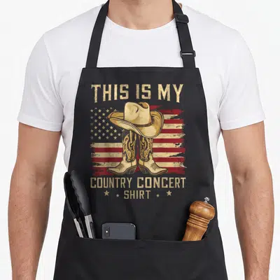 Funny concert country kitchen apron - Cowboy hat on boots before distressed flag with THIS IS MY COUNTRY CONCERT SHIRT — shown on a black cotton canvas apron against a white background