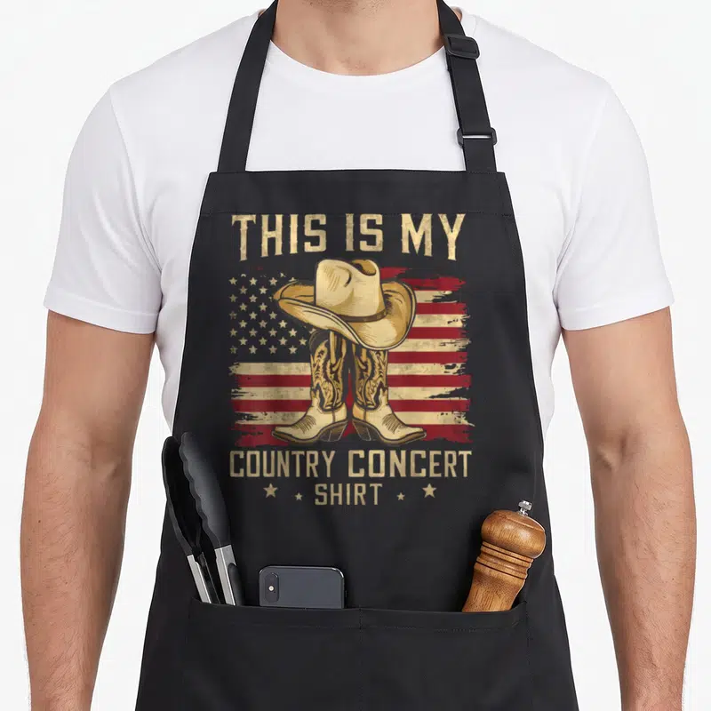 Funny concert country kitchen apron - Cowboy hat on boots before distressed flag with THIS IS MY COUNTRY CONCERT SHIRT — shown on a black cotton canvas apron against a white background