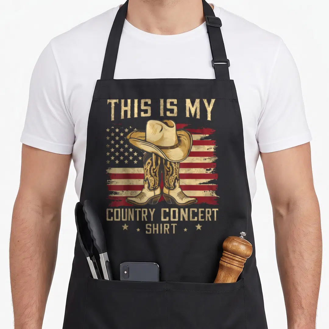 Funny concert country kitchen apron - Cowboy hat on boots before distressed flag with THIS IS MY COUNTRY CONCERT SHIRT — shown on a black cotton canvas apron against a white background