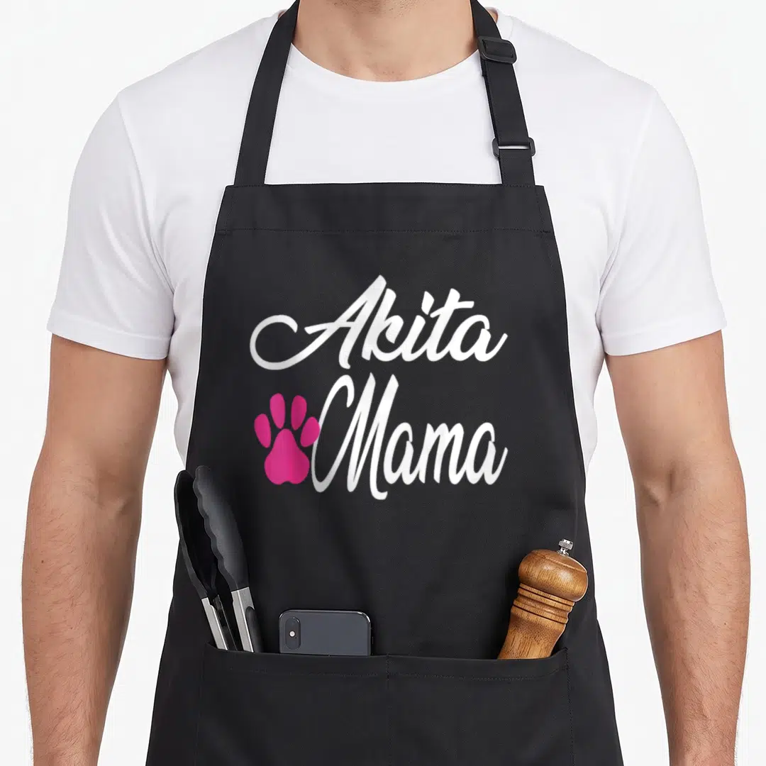 Cute akita dog apron - White Akita Mama text stacked above pink paw print on black background — shown on a black cotton canvas apron against a white background