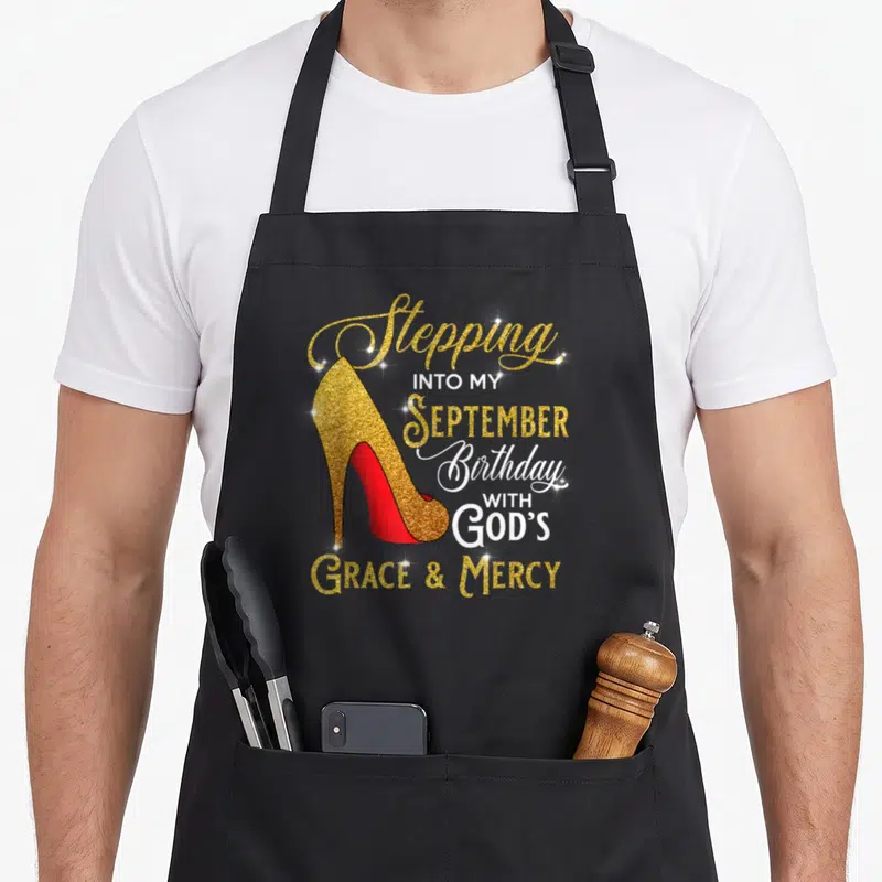September grace apron - Gold glitter high heel shoe with red sole and stacked birthday faith typography — shown on a black cotton canvas apron against a white background