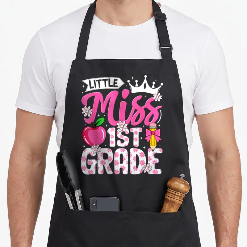 School cute grade apron - LITTLE Miss 1ST GRADE text with crown apple pencil bow floral fill pink white yellow green — shown on a black cotton canvas apron against a white background