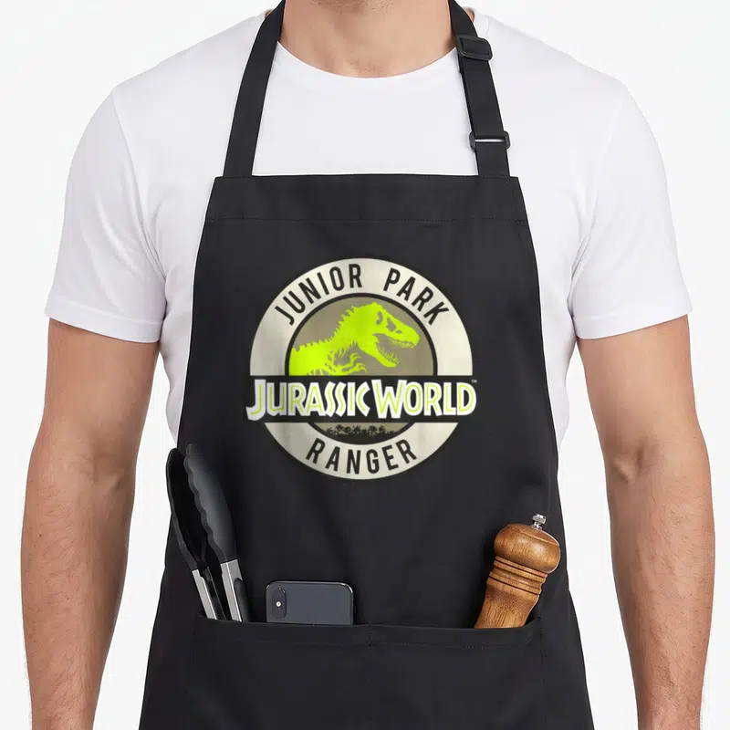Jurassic junior apron - Circular Jurassic World Junior Park Ranger logo with lime green dinosaur silhouette and palm — shown on a black cotton canvas apron against a white background