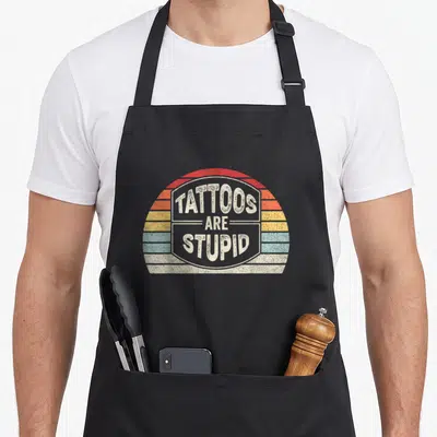 Catch tattoo apron - Retro text Tattoos Are Stupid over red orange yellow blue beige striped semi-circle — shown on a black cotton canvas apron against a white background