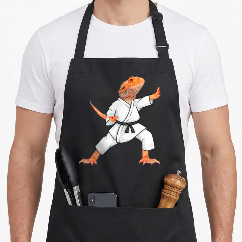 Doing dragon apron - Cartoon orange bearded dragon in white karate gi with black belt — shown on a black cotton canvas apron against a white background