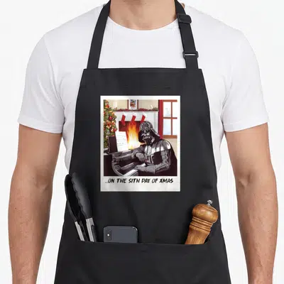 Sith xmas apron - Darth Vader plays piano before fireplace with stockings labeled Alin Luke Leia and Sith Day of Xmas — shown on a black cotton canvas apron against a white background