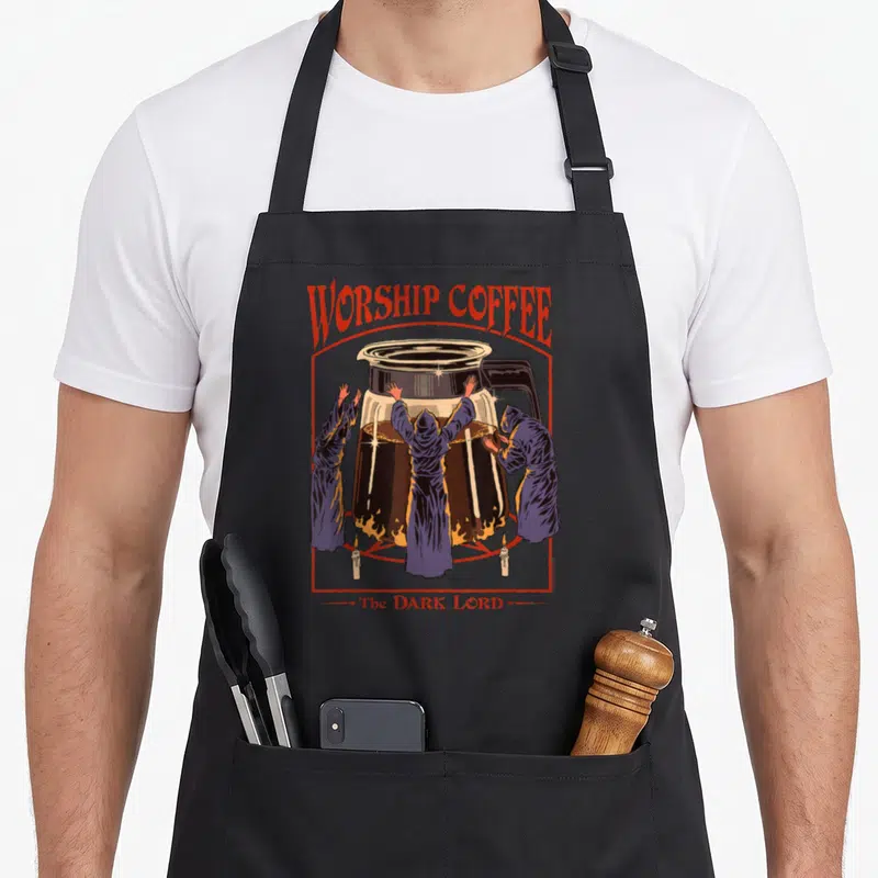 Steven coffee apron - Retro graphic of hooded figures worshipping a coffee pot with WORSHIP COFFEE text — shown on a black cotton canvas apron against a white background