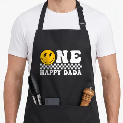Dada family cooking aprons - ONE HAPPY DADA text with yellow lightning eye smiley face and checkered strip — shown on a black cotton canvas apron against a white background