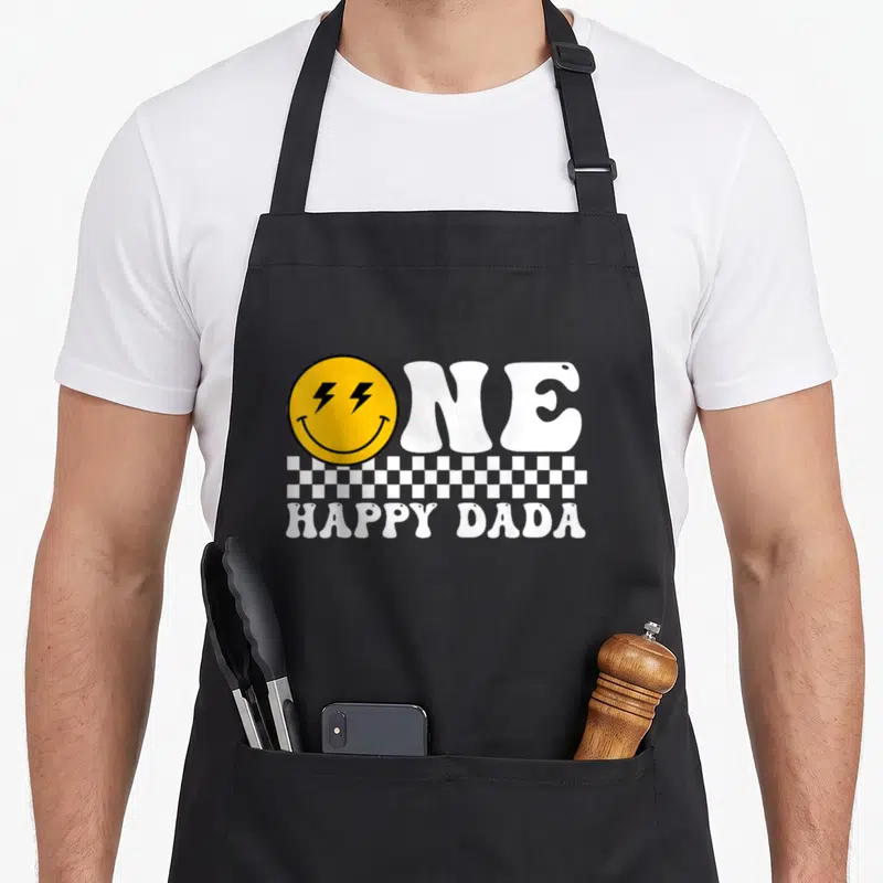 Dada family cooking aprons - ONE HAPPY DADA text with yellow lightning eye smiley face and checkered strip — shown on a black cotton canvas apron against a white background