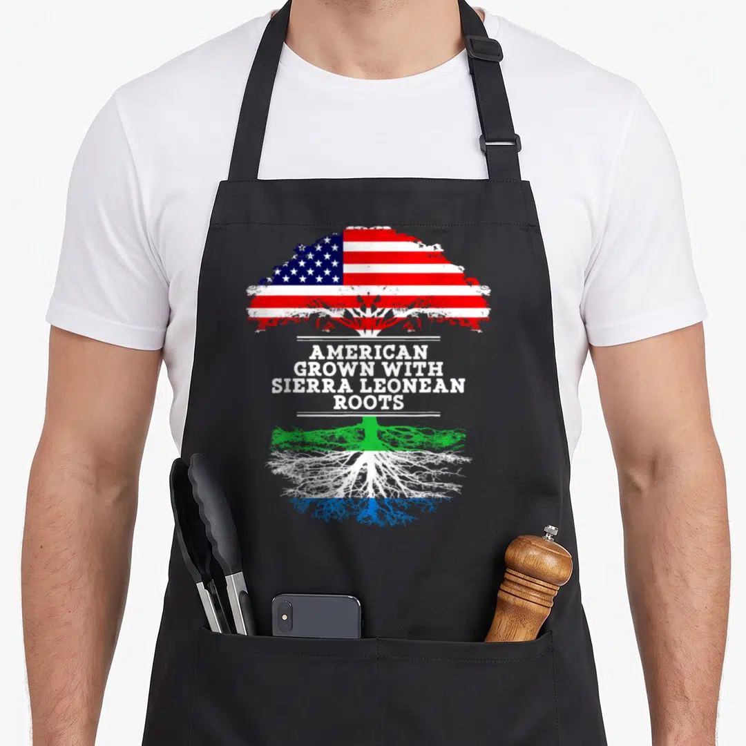 Leonean root sierra apron - Tree with US flag canopy and green white blue roots surrounding stacked text — shown on a black cotton canvas apron against a white background