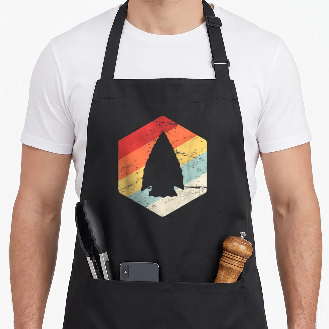 Artifact hunter apron - Grey arrowhead silhouette inside a hexagon filled with horizontal red orange yellow blue — shown on a black cotton canvas apron against a white background