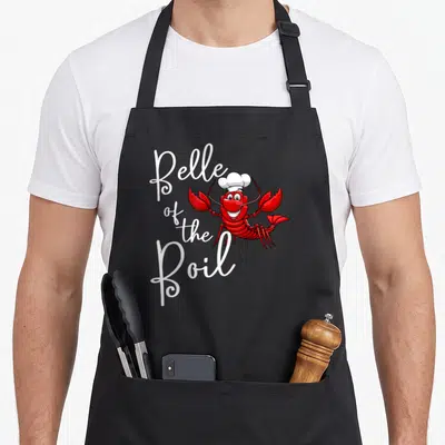 Belle lobster apron - Red cartoon lobster in chef hat next to white Belle of the Boil script text — shown on a black cotton canvas apron against a white background