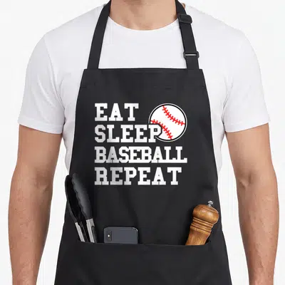 Sleep baseball apron - White EAT SLEEP BASEBALL REPEAT text with red stitched ball overlapping SLEEP — shown on a black cotton canvas apron against a white background