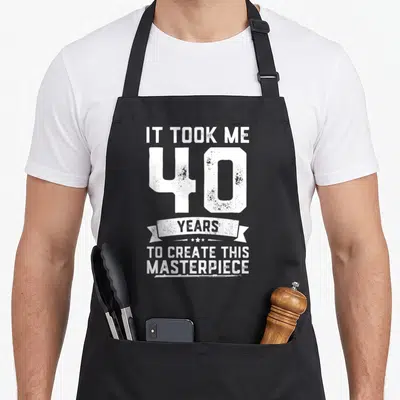Gag 40th apron - Distressed white text It Took Me 40 Years to Create This Masterpiece with ribbon and stars — shown on a black cotton canvas apron against a white background