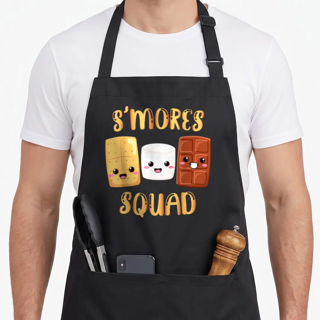Squad camping apron - Cartoon graham cracker, marshmallow, and chocolate bar with smiling faces and orange text — shown on a black cotton canvas apron against a white background