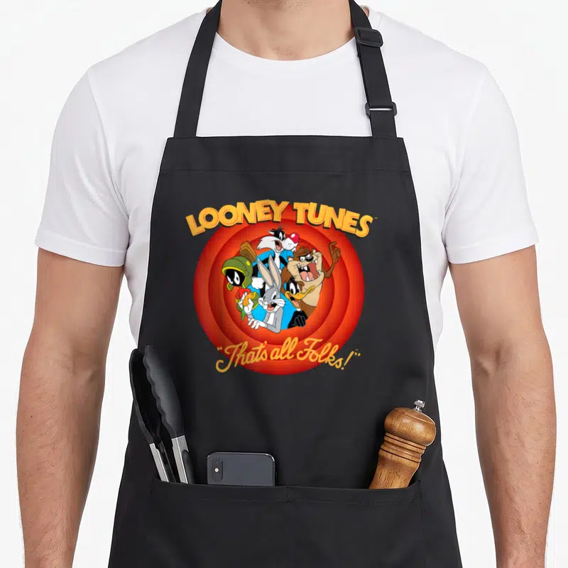 Funny folk apron - Looney Tunes characters clustered over red orange circles with arched text and cursive quote below — shown on a black cotton canvas apron against a white background