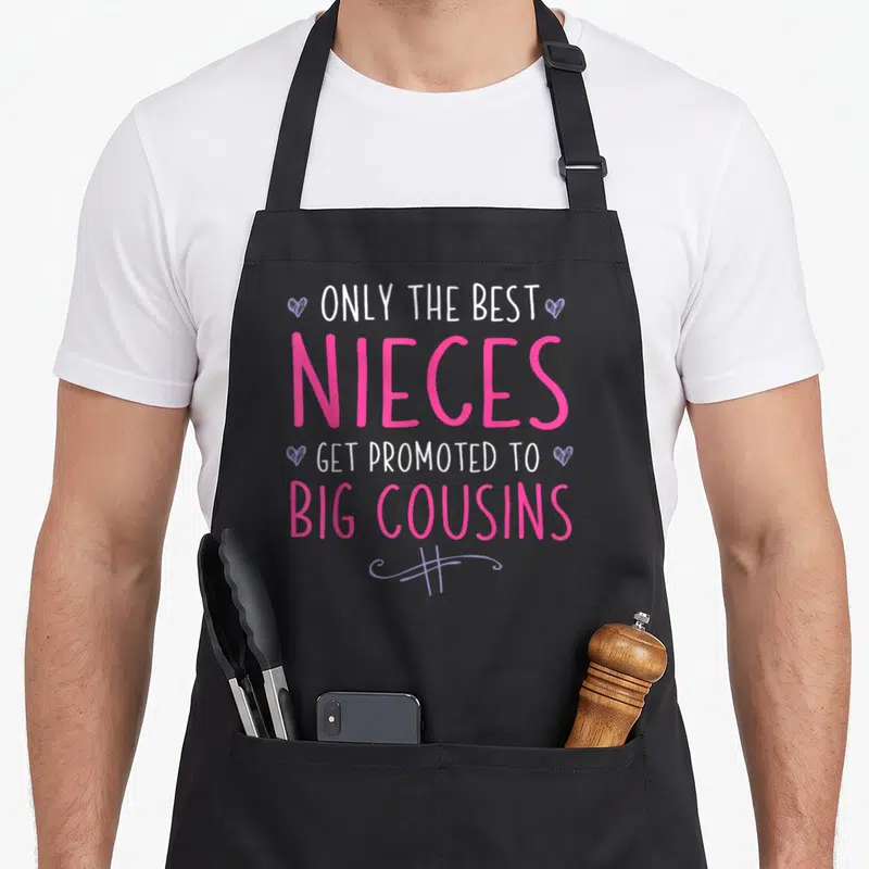 Niece birthday surprise apron - Stacked white and pink text reading ONLY THE BEST NIECES GET PROMOTED TO BIG COUSINS — shown on a black cotton canvas apron against a white background