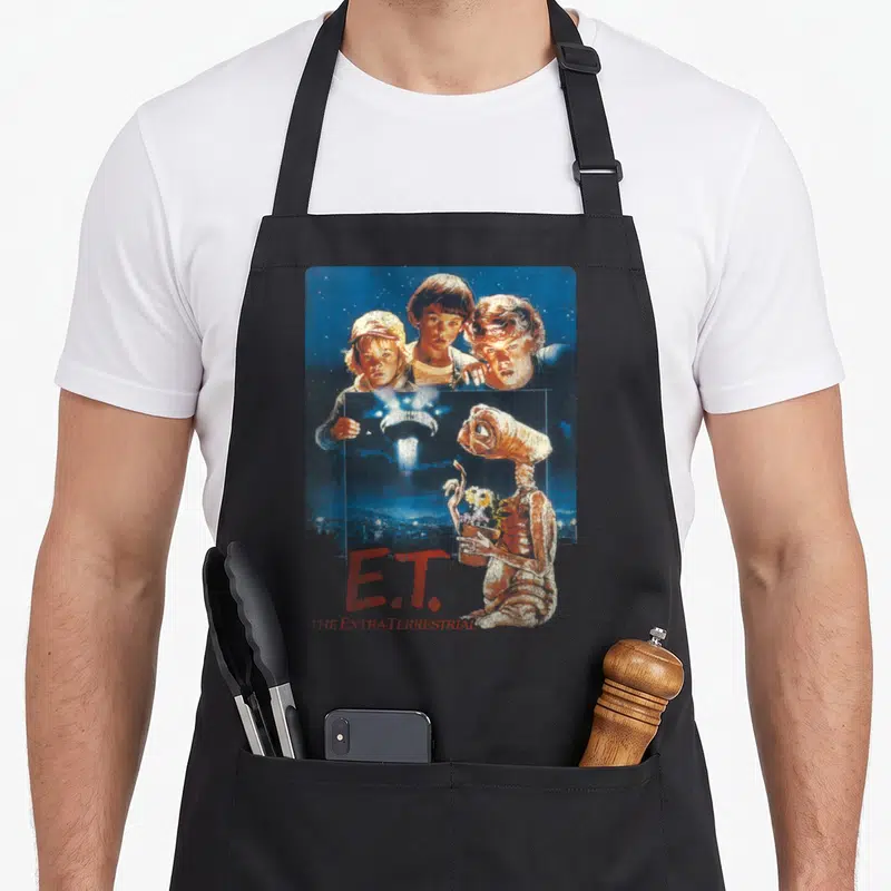 Photograph e.a apron - E.T. holding flowers, three boys, spaceship, red text on blue background — shown on a black cotton canvas apron against a white background