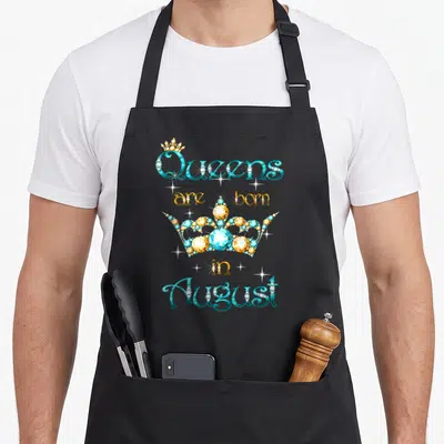 Cute august queen apron - Stacked teal and gold text reading Queens are born in August with a large jeweled crown — shown on a black cotton canvas apron against a white background