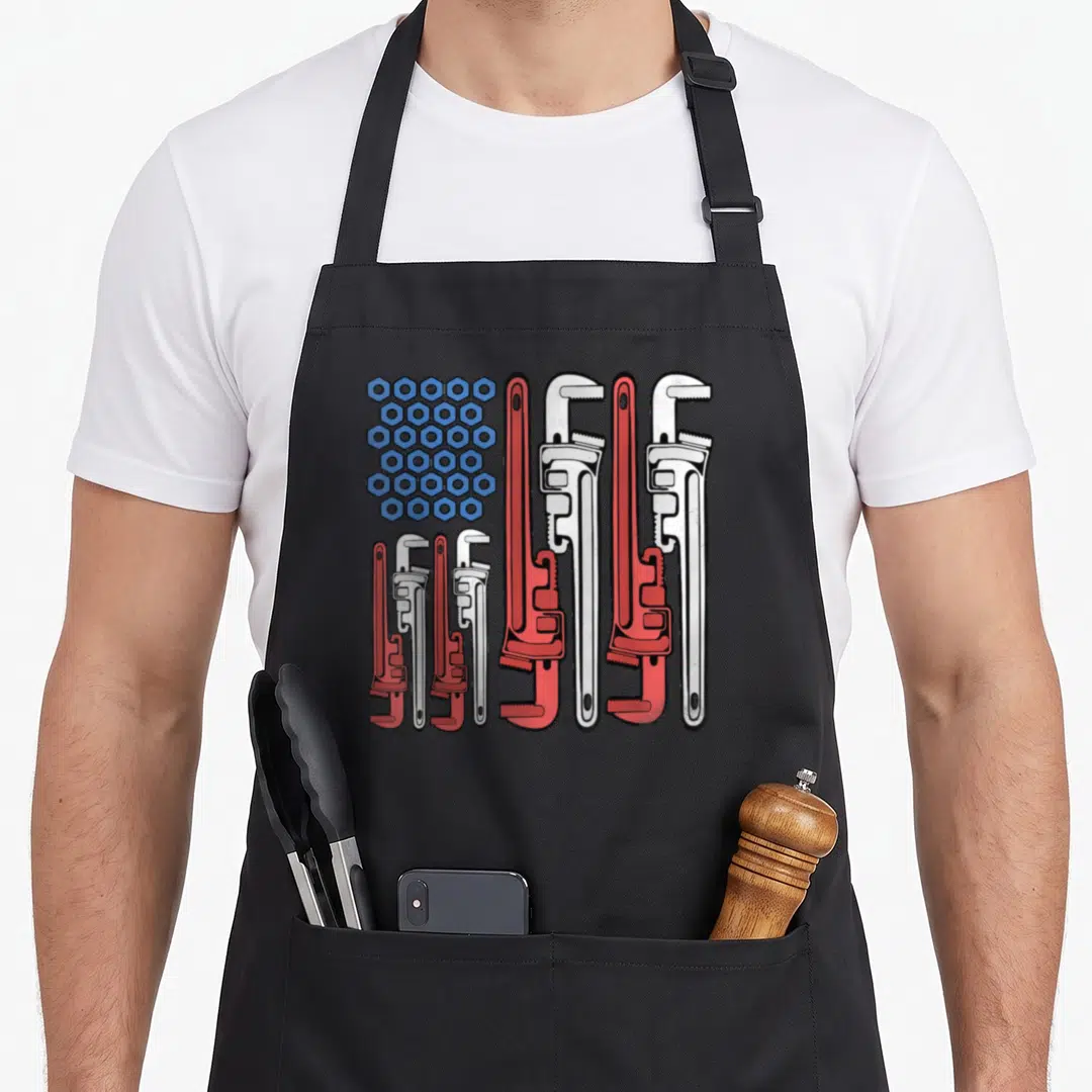 Pipe 4th of july apron - Blue nuts and red white pipe wrenches forming a cartoon American flag pattern — shown on a black cotton canvas apron against a white background