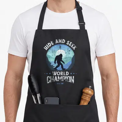 Funny seek bigfoot apron - Bigfoot silhouette in blue mountain circle with hide and seek world champion text — shown on a black cotton canvas apron against a white background