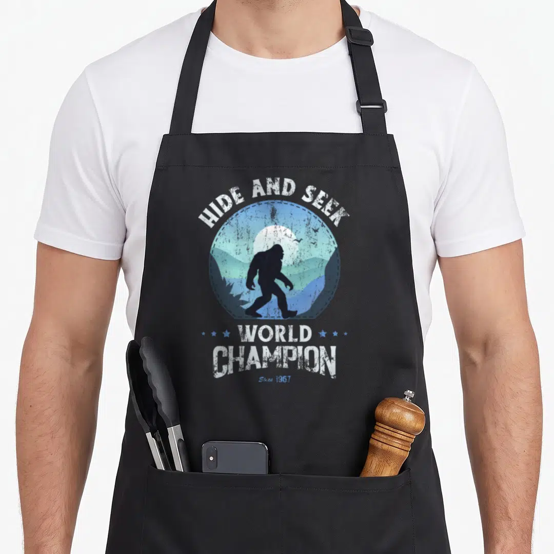 Funny seek bigfoot apron - Bigfoot silhouette in blue mountain circle with hide and seek world champion text — shown on a black cotton canvas apron against a white background