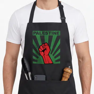 Propaganda palestine apron - Red raised fist on green sunburst rays with PALESTINE text in distressed style — shown on a black cotton canvas apron against a white background
