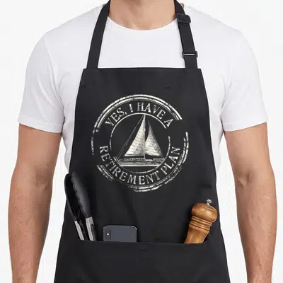 Sailing plain apron - Distressed circular graphic with sailboat and text Yes I Have a Retirement Plan in off-white — shown on a black cotton canvas apron against a white background