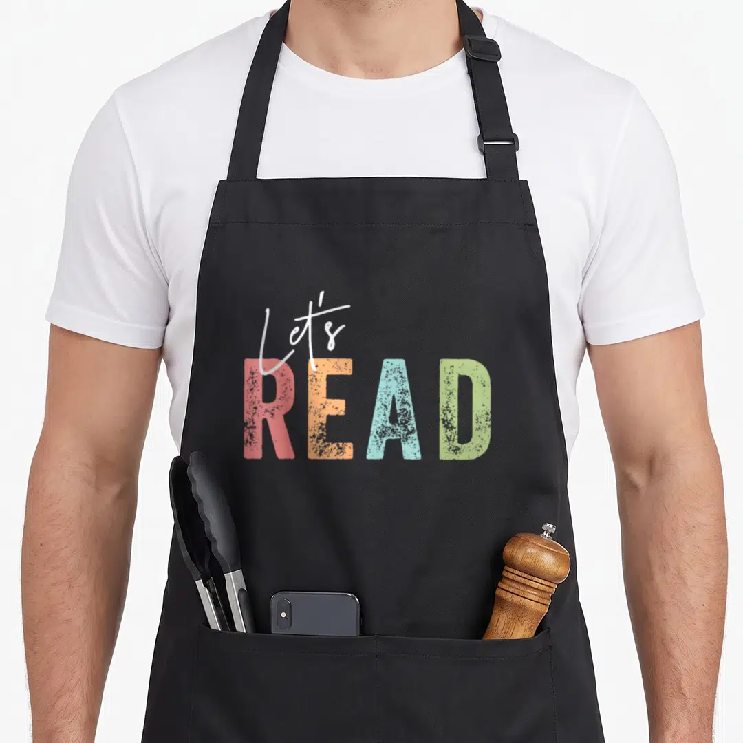 Librarian teacher apron - Let's READ text in pink orange blue and green stacked typography — shown on a black cotton canvas apron against a white background