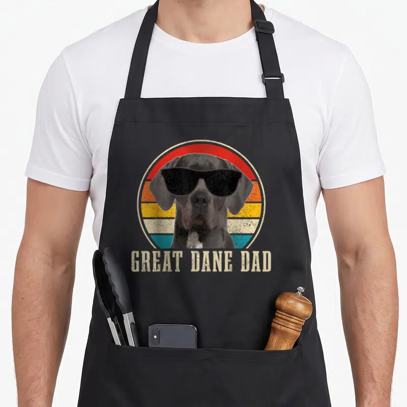 Danish dane dog apron - Retro Great Dane in sunglasses before colored stripes with text GREAT DANE DAD below — shown on a black cotton canvas apron against a white background