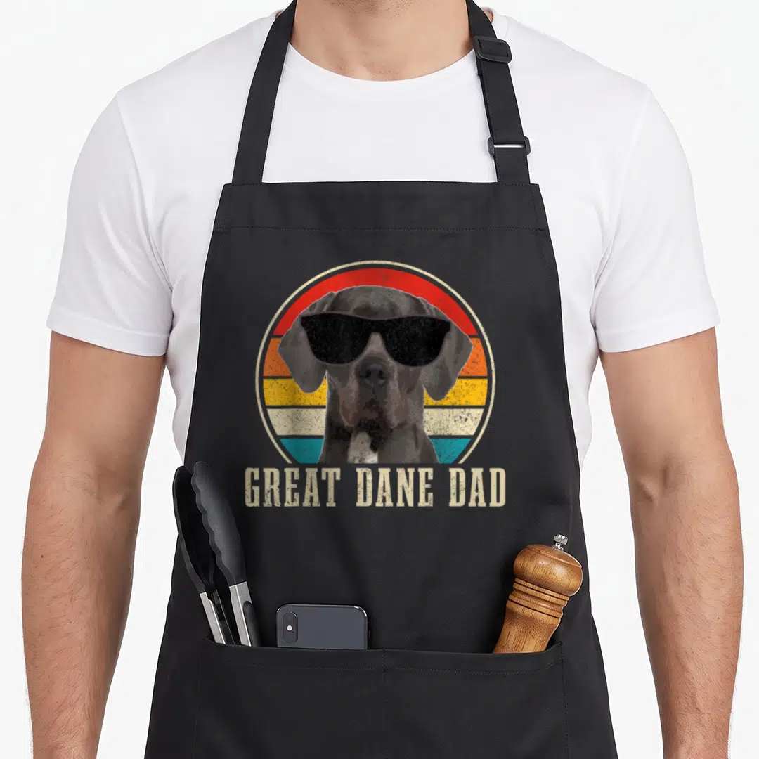 Danish dane dog apron - Retro Great Dane in sunglasses before colored stripes with text GREAT DANE DAD below — shown on a black cotton canvas apron against a white background