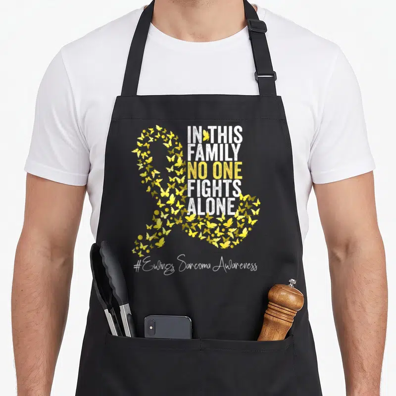 Ewing ribbon apron - Yellow butterfly ribbon shape with In This Family No One Fights Alone text and hashtag — shown on a black cotton canvas apron against a white background
