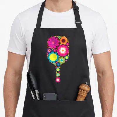 Sport pickleball apron - Pickleball paddle shape formed by pink, orange, yellow, blue, and green cartoon flowers — shown on a black cotton canvas apron against a white background
