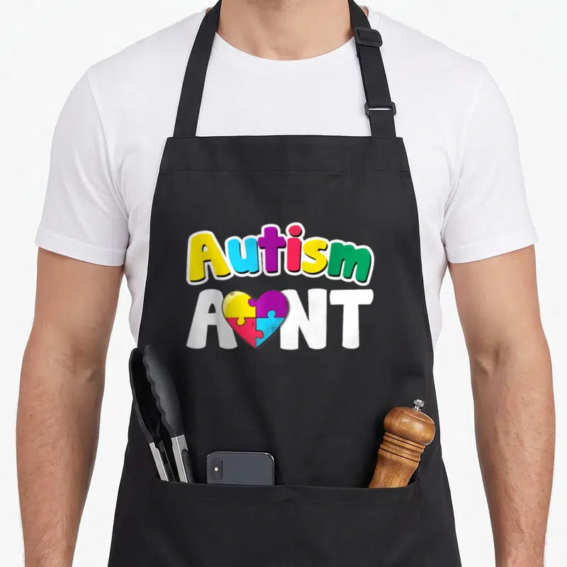 Aunt puzzles apron - Autism A NT text with puzzle heart in yellow blue purple pink green white — shown on a black cotton canvas apron against a white background