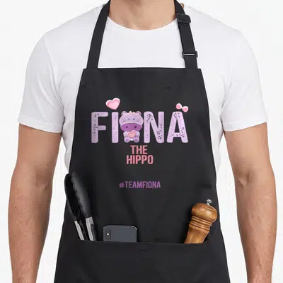 Hippo fiona apron - Cartoon purple hippo inside FIONA lettering with pink hearts and bow, #TEAMFIONA below — shown on a black cotton canvas apron against a white background