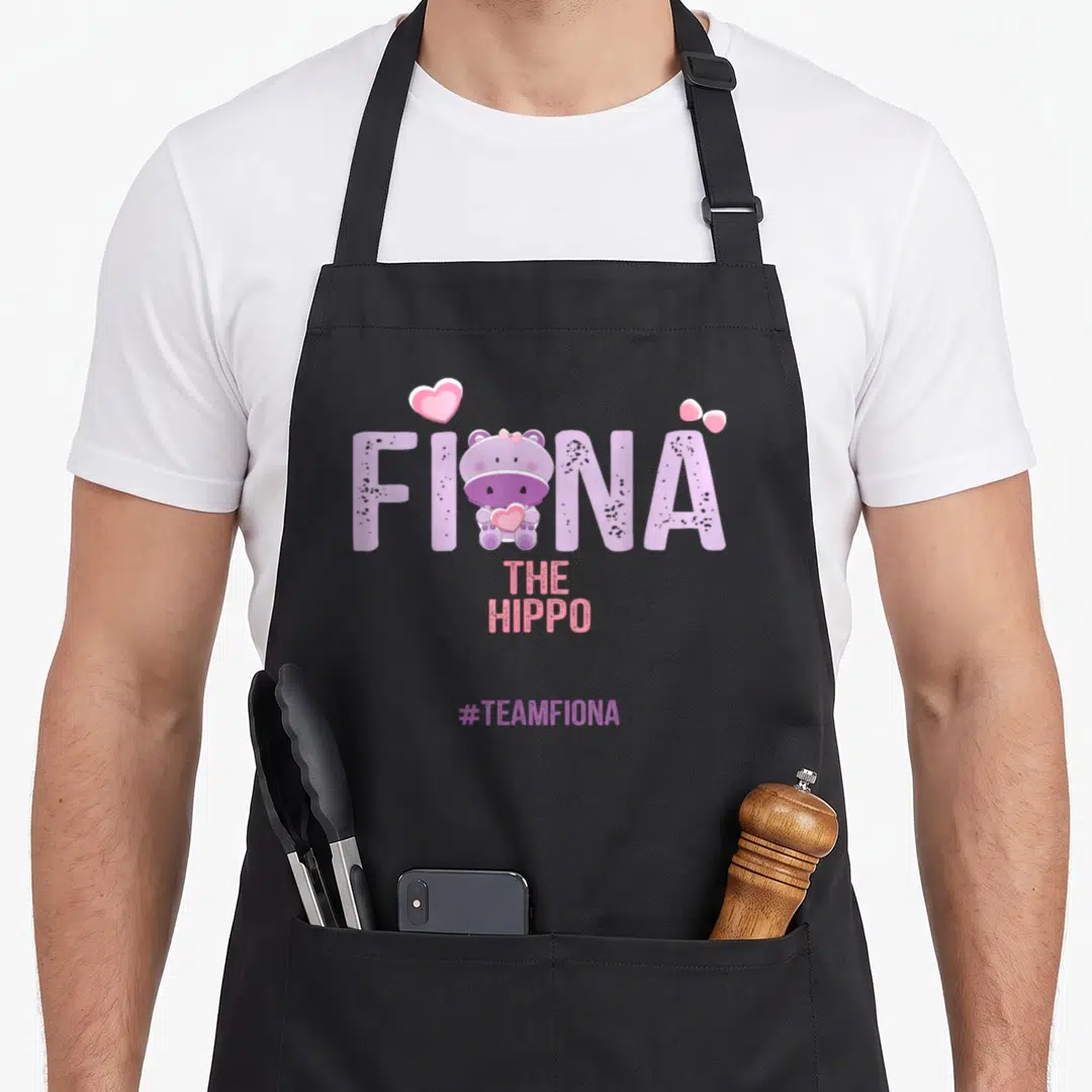 Hippo fiona apron - Cartoon purple hippo inside FIONA lettering with pink hearts and bow, #TEAMFIONA below — shown on a black cotton canvas apron against a white background