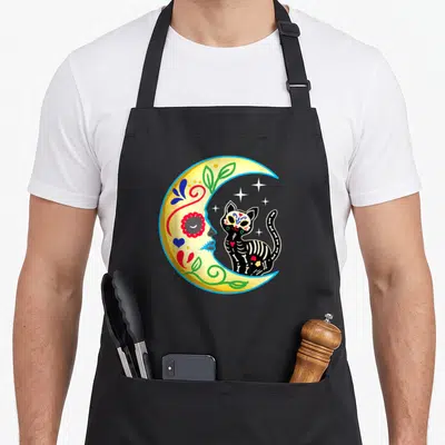 Cat day of the dead apron - Cartoon skeleton cat sitting in a decorated yellow crescent moon with stars — shown on a black cotton canvas apron against a white background