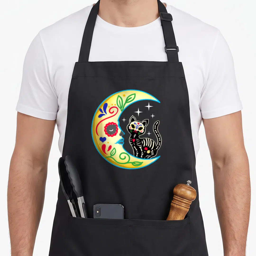 Cat day of the dead apron - Cartoon skeleton cat sitting in a decorated yellow crescent moon with stars — shown on a black cotton canvas apron against a white background