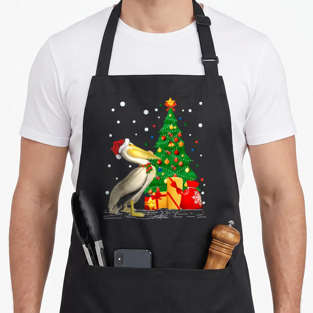 Pelican christmas santa apron - Cartoon pelican in red hat beside Christmas tree with gifts and snowflakes — shown on a black cotton canvas apron against a white background