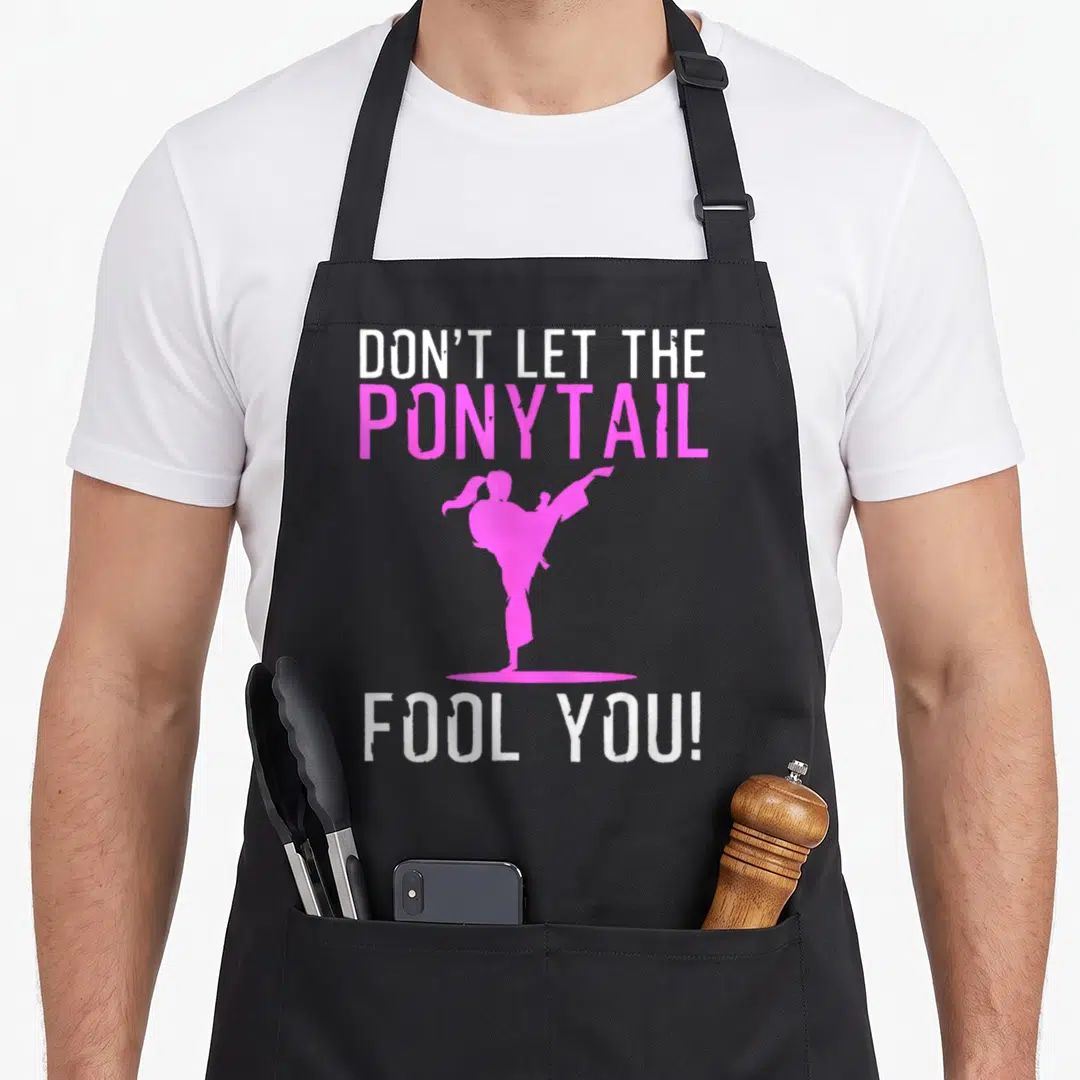 Kick karate apron - Pink martial artist kicking silhouette between DON'T LET THE PONYTAIL and FOOL YOU! text — shown on a black cotton canvas apron against a white background