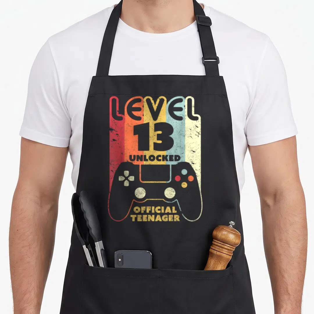Teenage birthday apron - Level 13 Unlocked game controller silhouette over red orange yellow blue grey vertical stripes — shown on a black cotton canvas apron against a white background