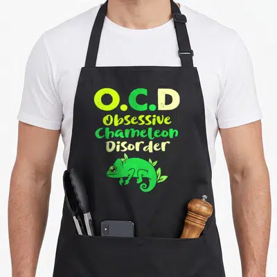 Obsessive chameleon apron - O.C.D text stacked above a green cartoon chameleon with leaves — shown on a black cotton canvas apron against a white background