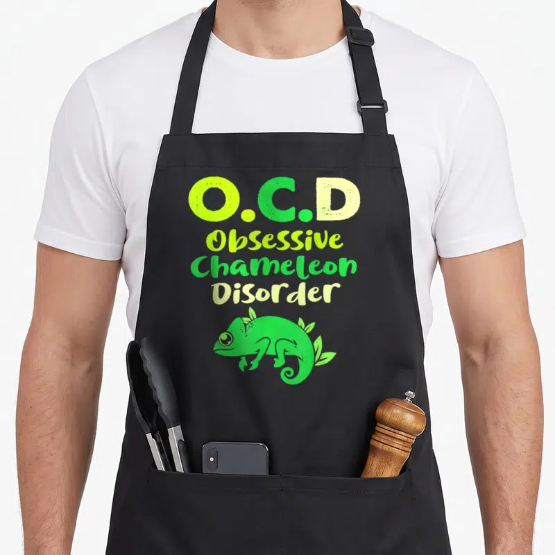 Obsessive chameleon apron - O.C.D text stacked above a green cartoon chameleon with leaves — shown on a black cotton canvas apron against a white background