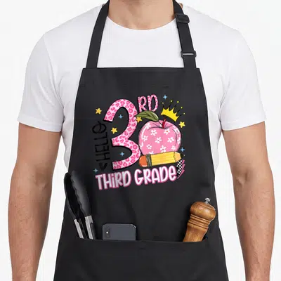 3rd third teacher apron - Pink leopard number 3 with floral apple, pencil, crown, and HELLO THIRD GRADE text — shown on a black cotton canvas apron against a white background
